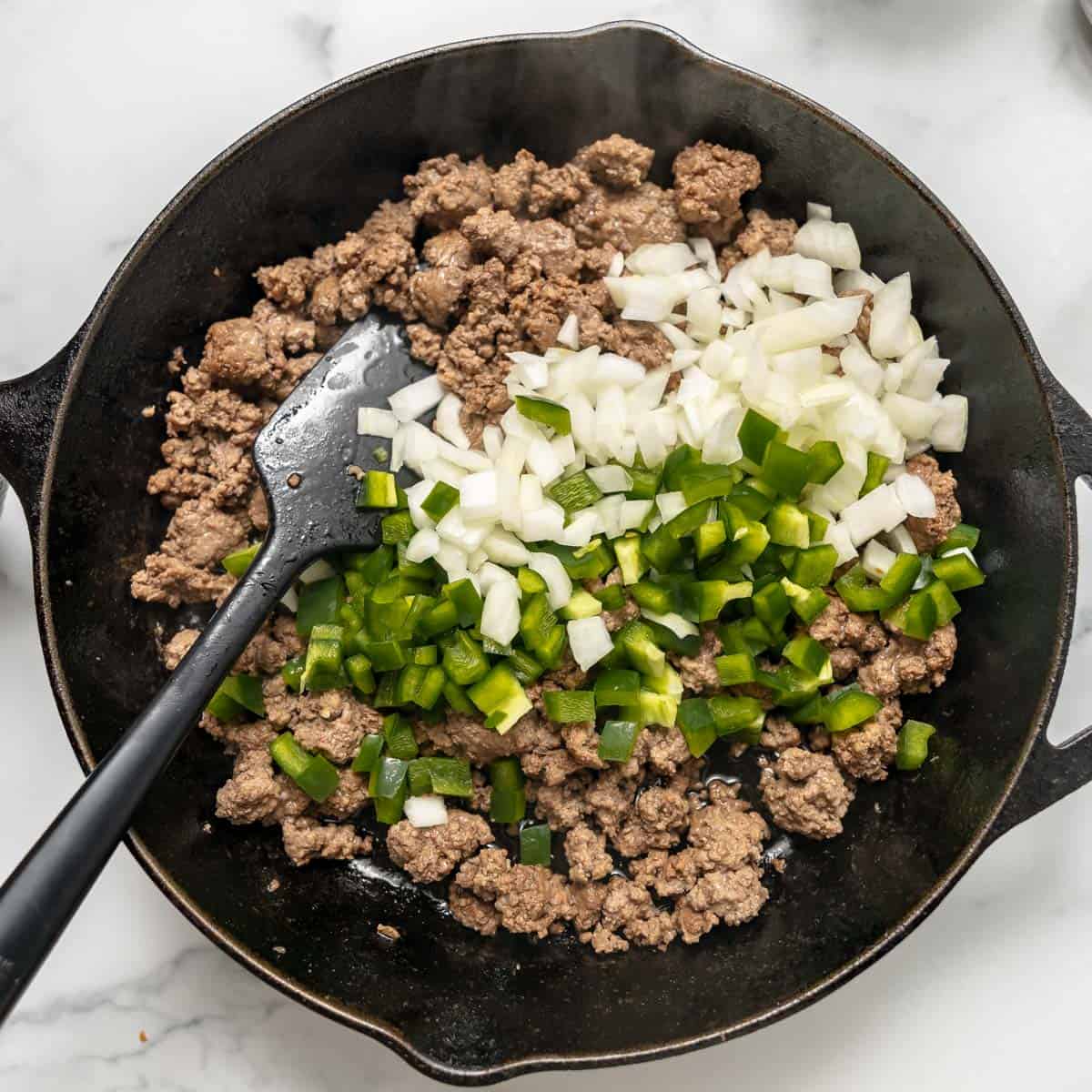 Ground beef, diced onions, and chopped green bell peppers cooking in a black skillet with a spatula on a marble surface.