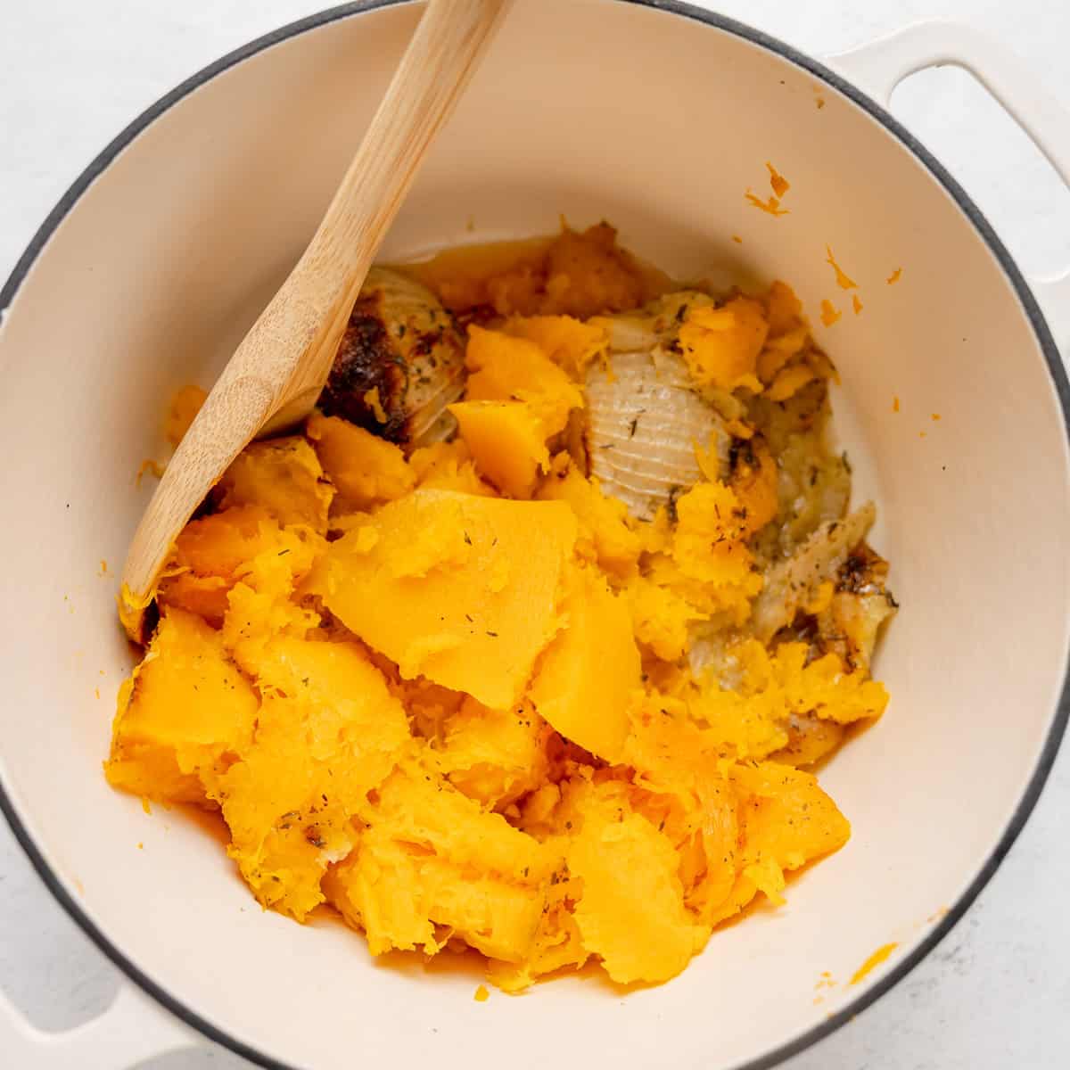 A white pot with cooked, mashed butternut squash and onion, stirred with a wooden spoon.