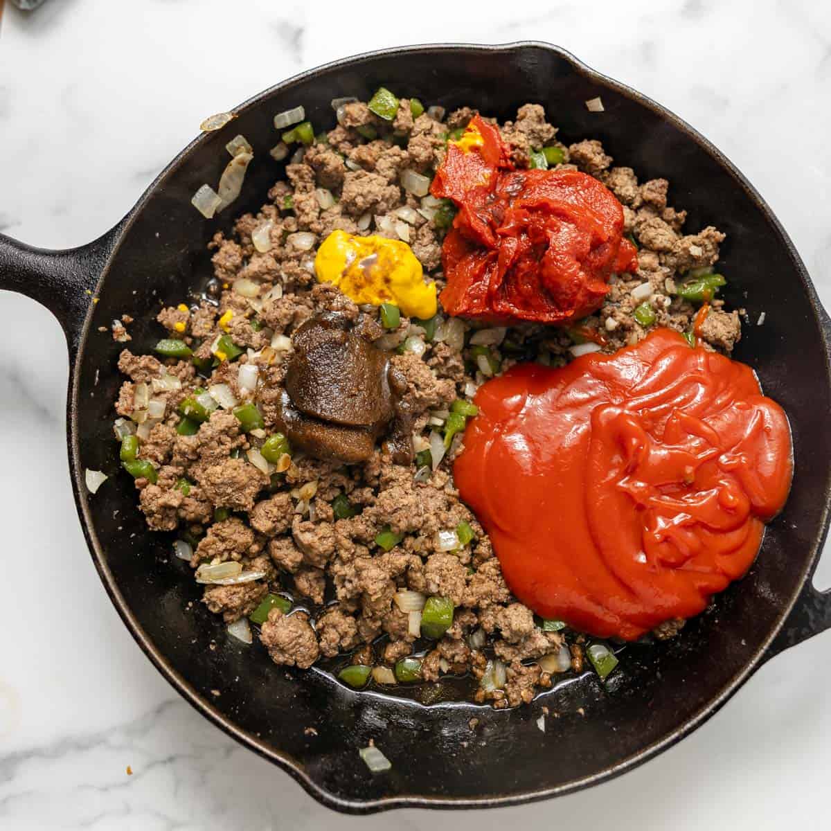 A cast iron skillet with cooked ground beef, onions, and peppers, topped with ketchup, tomato paste, mustard, and a brown paste, on a marble surface.