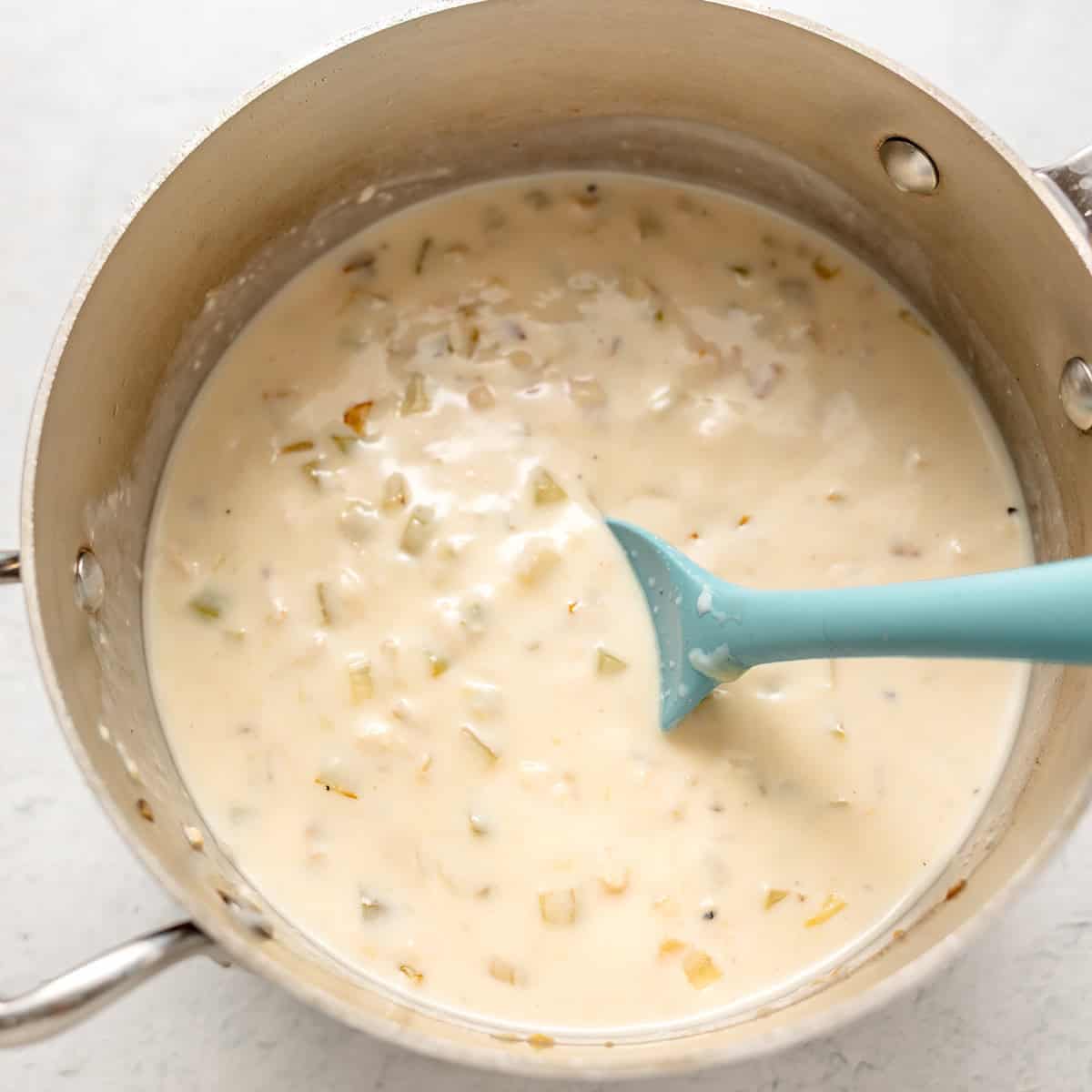 A metal saucepan filled with a creamy white sauce containing chopped vegetables, being stirred with a blue silicone spoon.
