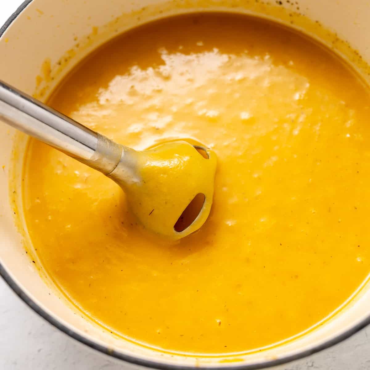 An immersion blender sits in a pot of smooth, orange soup, partially submerged and coated with the blended soup mixture.