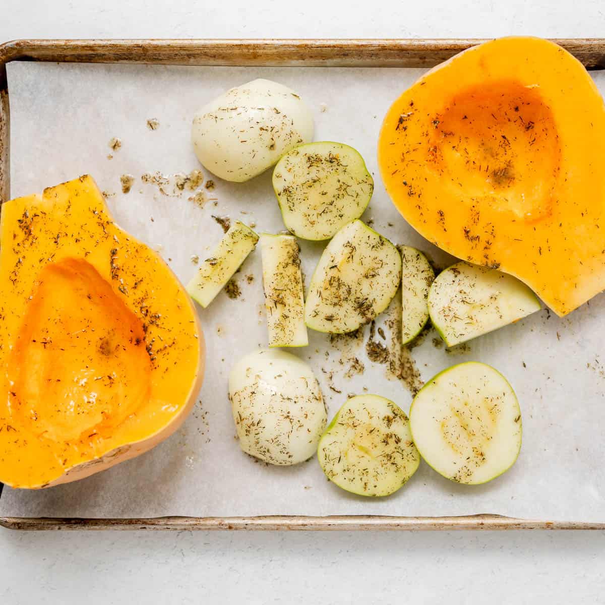 Halved butternut squash and sliced apples and onions on a parchment-lined baking sheet, sprinkled with herbs and seasoning, ready for roasting.
