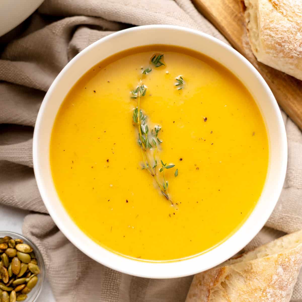 A white bowl filled with creamy orange soup, garnished with fresh thyme, sits on a beige cloth next to bread and a small dish of pumpkin seeds.