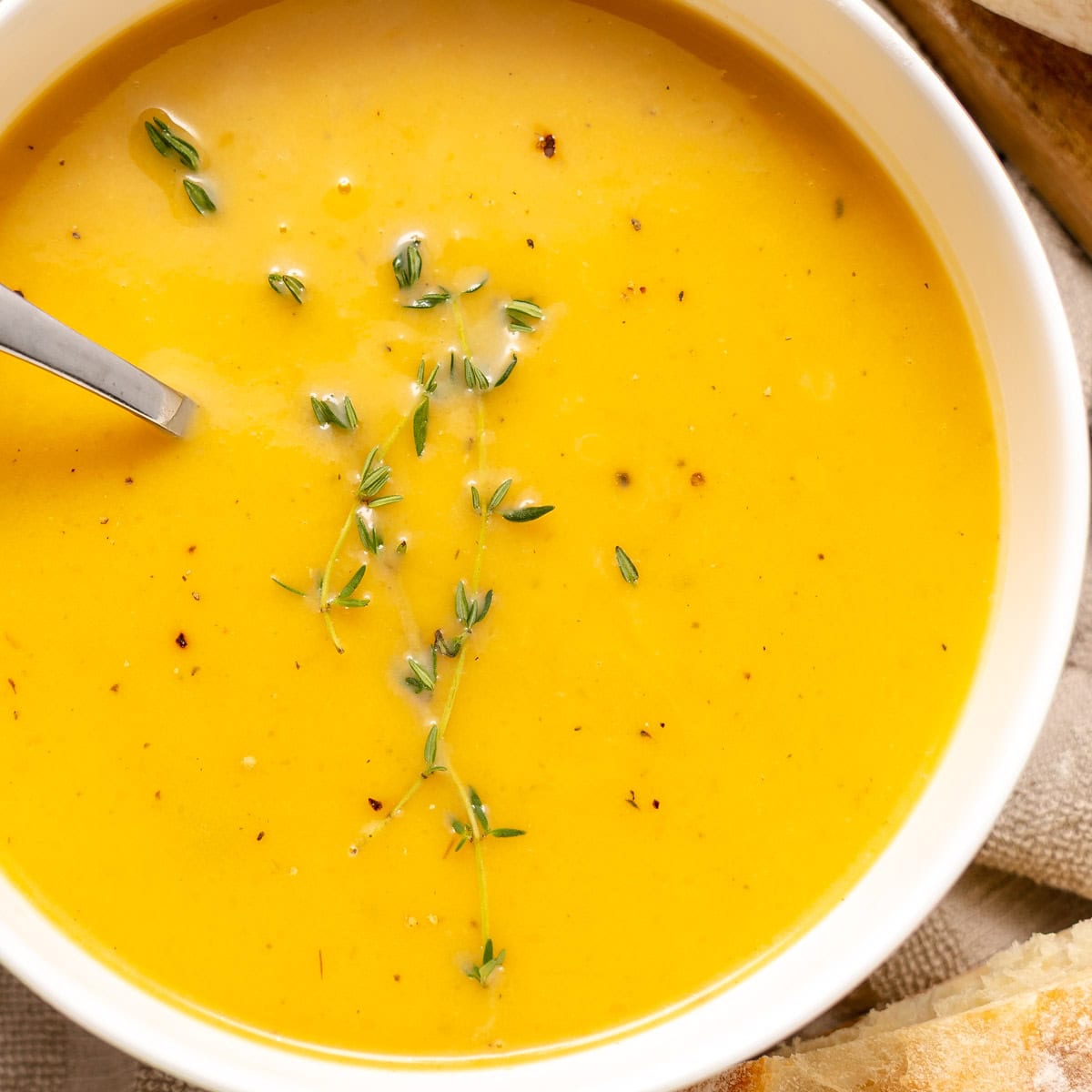A bowl of creamy orange soup garnished with fresh thyme, with a spoon in the bowl and a slice of bread partially visible on the side.