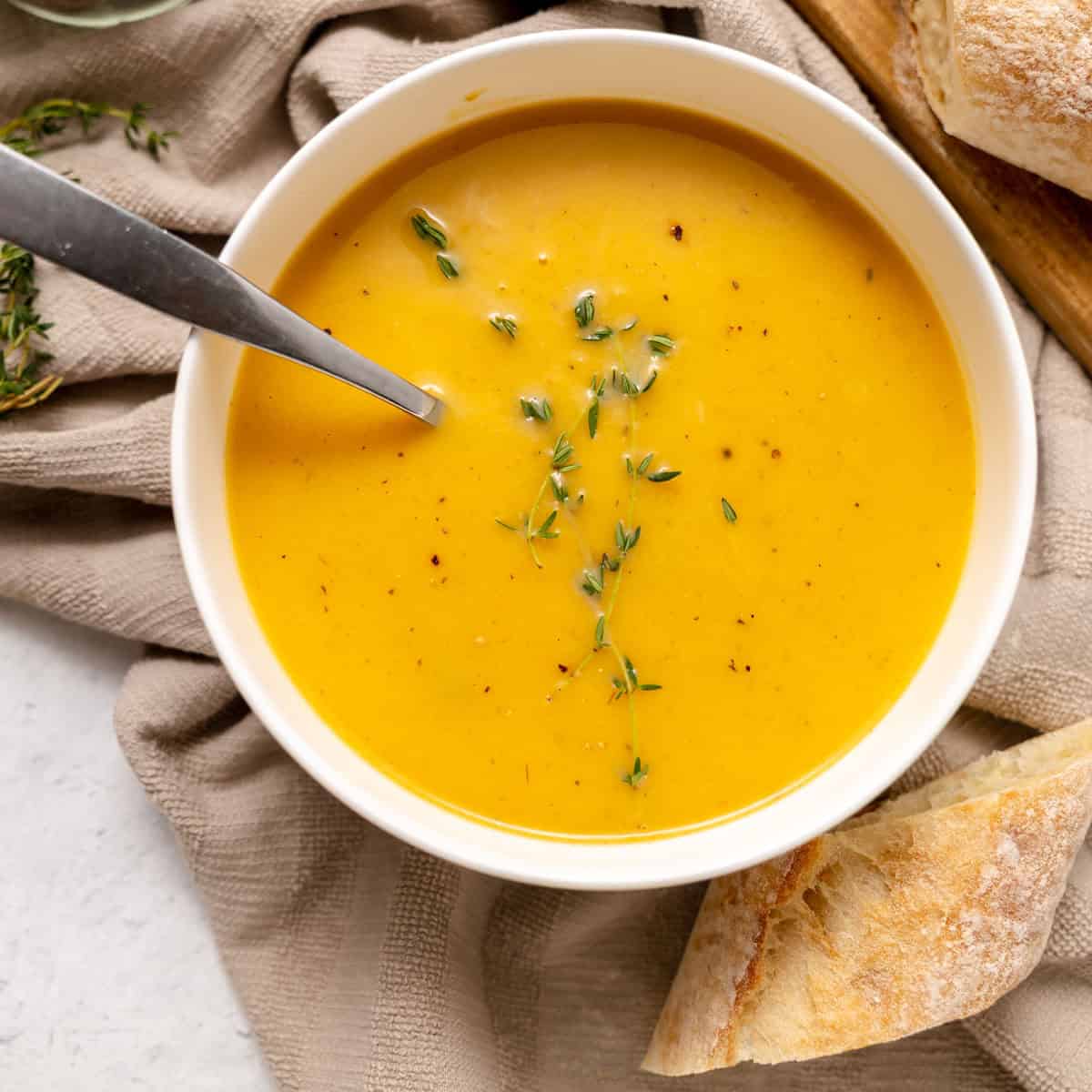 Bowl of creamy butternut squash soup garnished with herbs, with a spoon inside and pieces of bread on the side.