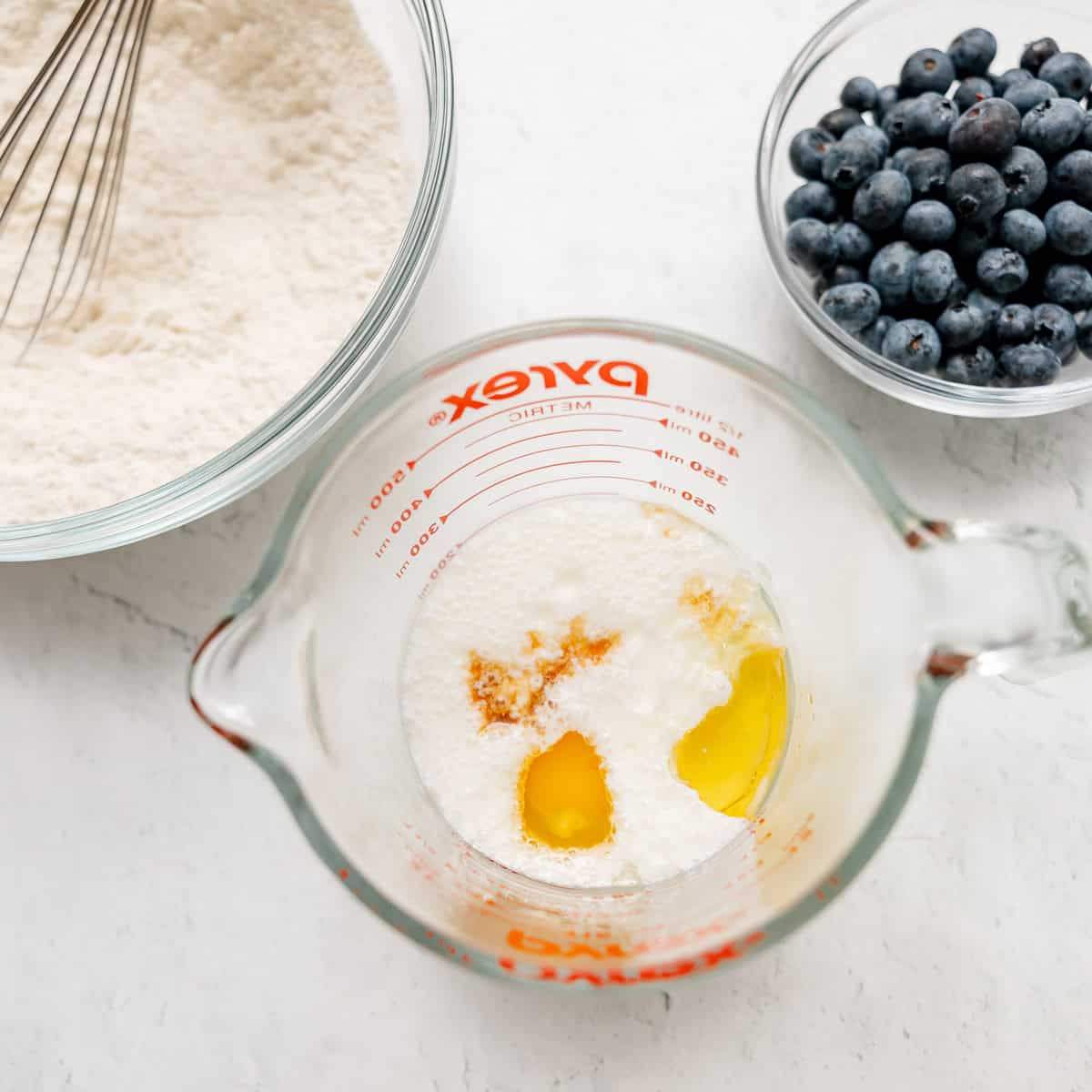 A glass bowl of flour, a bowl of blueberries, and a measuring cup with milk, eggs, and vanilla on a white surface.