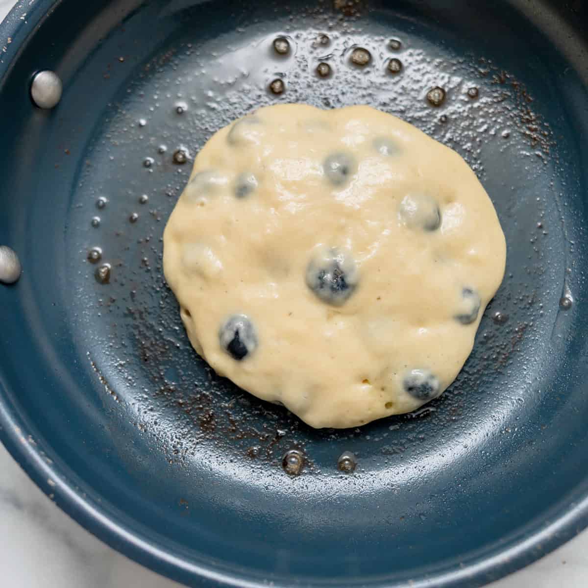 Uncooked blueberry pancake batter in a nonstick skillet with small droplets of oil visible around the batter.