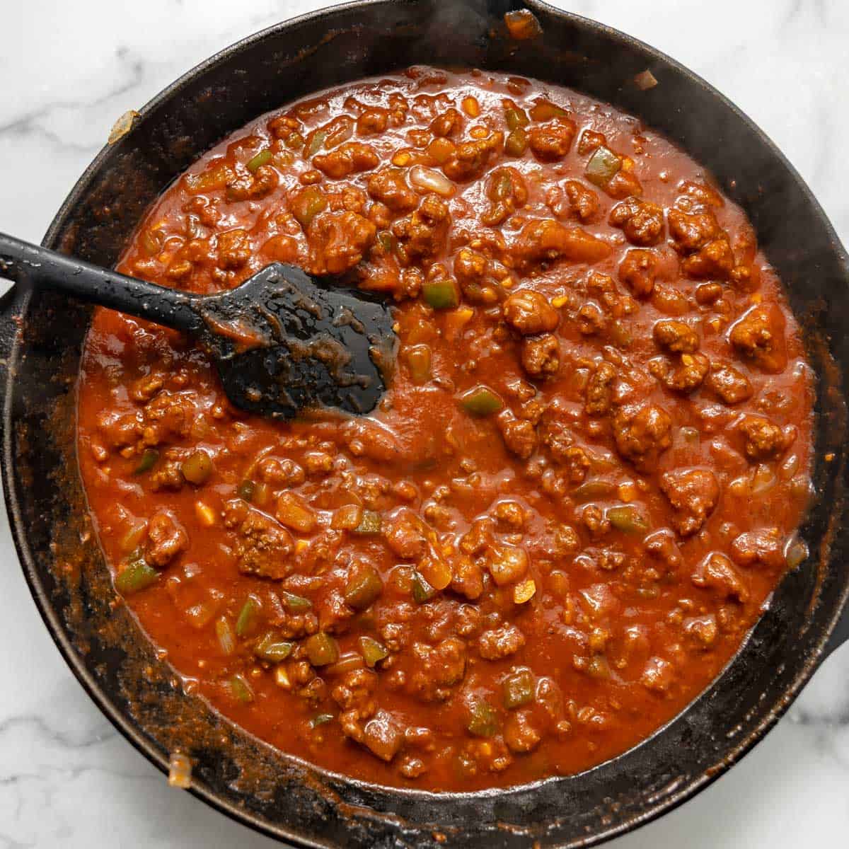 A skillet of thick, chunky tomato-based meat sauce with green peppers being stirred with a black spatula.