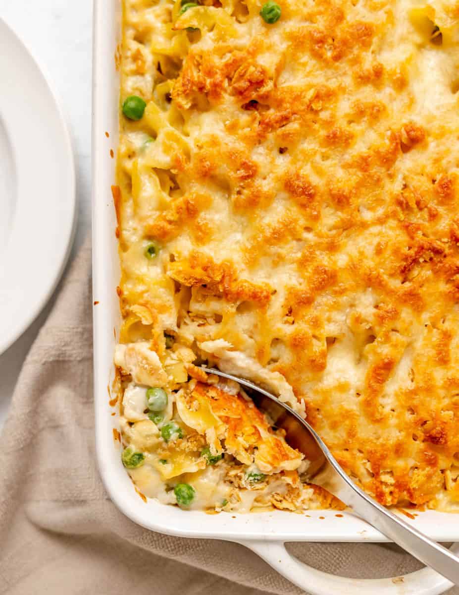 Baked casserole with melted cheese on top, showing noodles, peas, and pieces of tuna, with a serving spoon scooping out a portion.