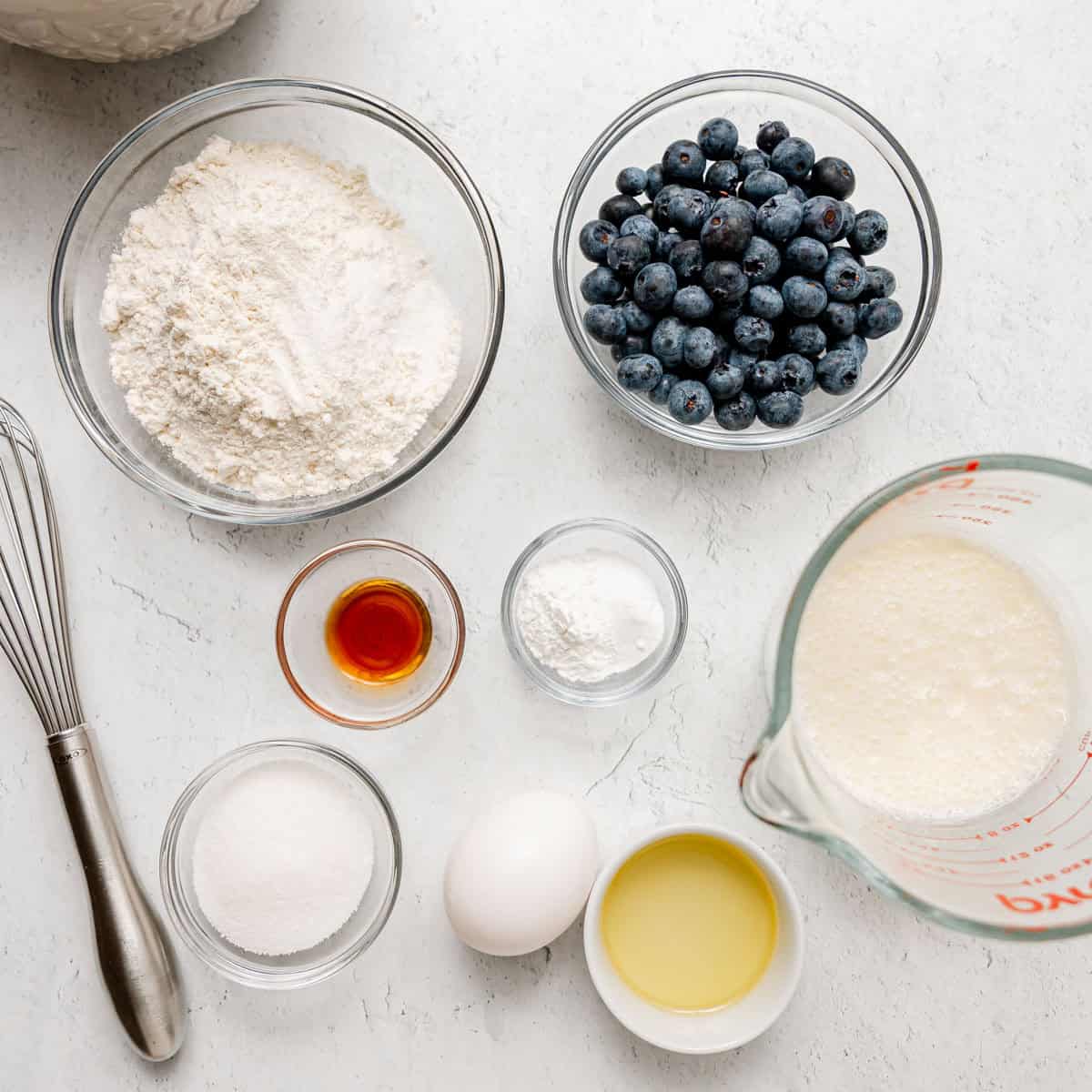 Bowls with flour, blueberries, sugar, baking powder, vanilla, oil, an egg, and a measuring cup of milk are arranged on a white countertop next to a whisk.