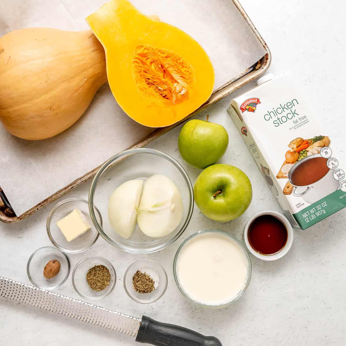 Halved butternut squash, two green apples, onion, carton of chicken stock, cream, butter, spices, and a grater arranged on a baking sheet.