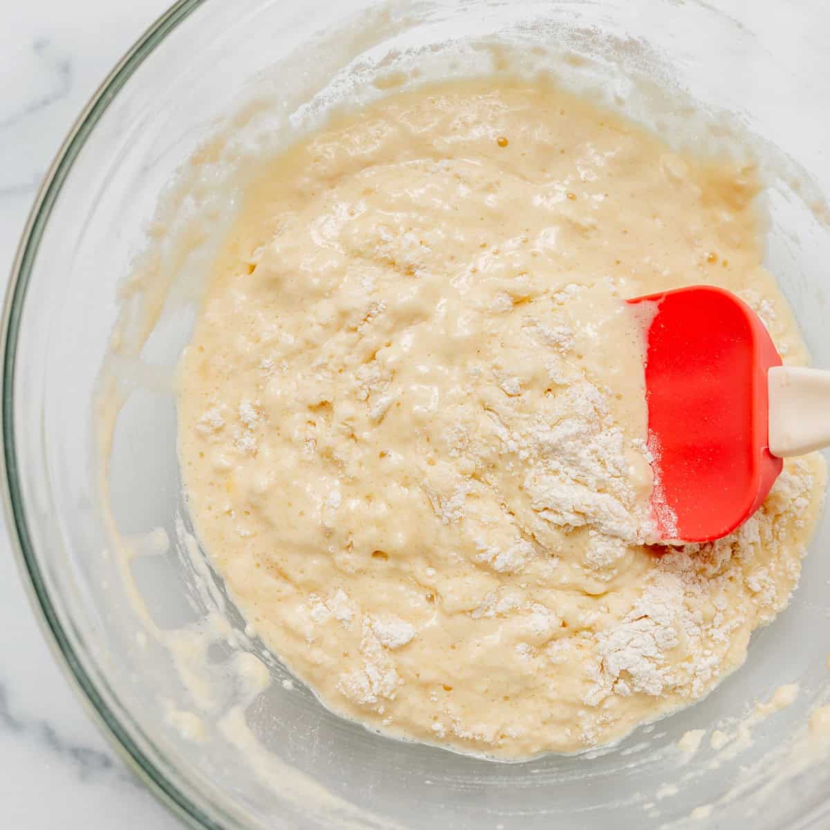 Bowl of pancake batter being mixed with a red spatula on a white surface.