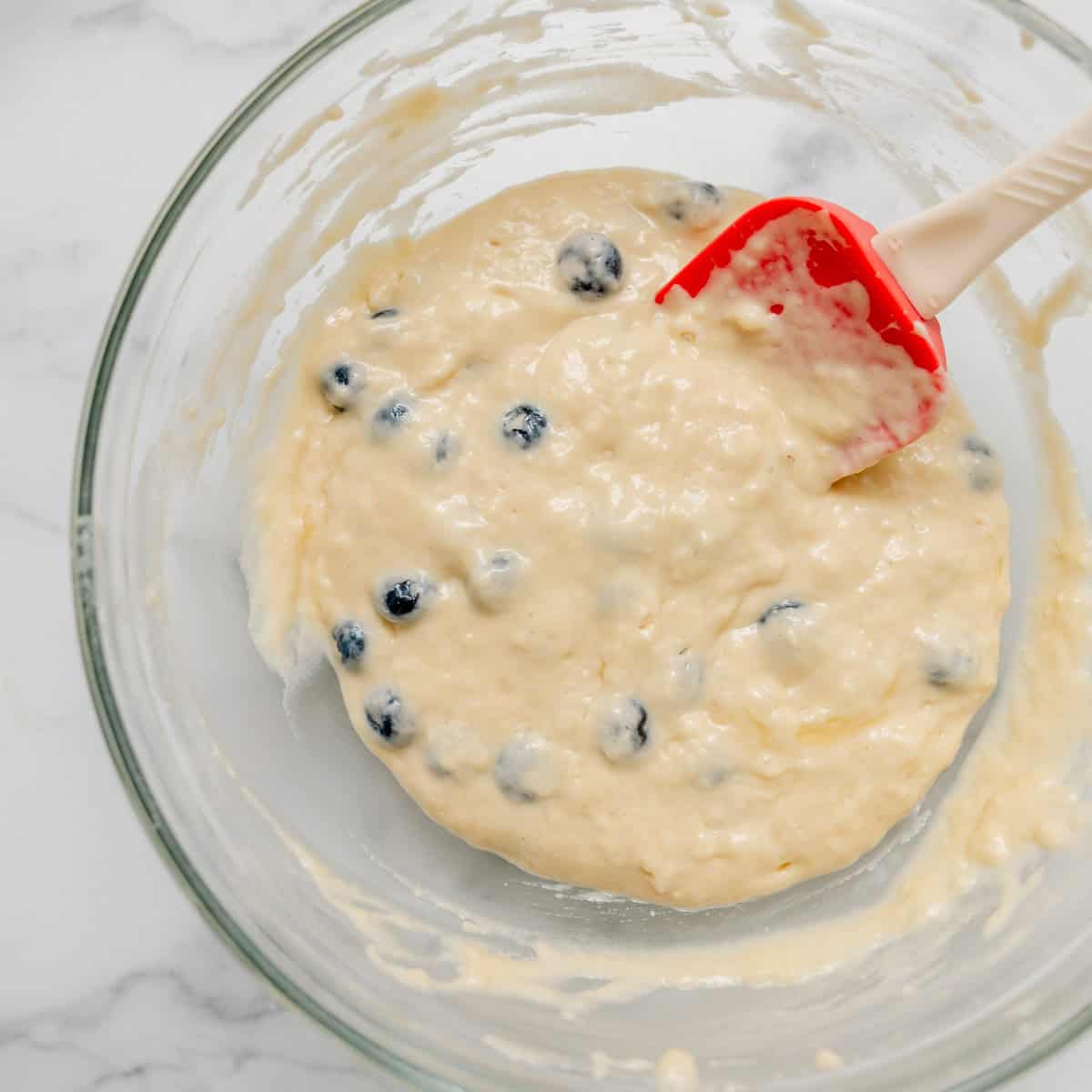 A glass bowl filled with blueberry pancake batter being mixed with a red and white spatula on a white surface.