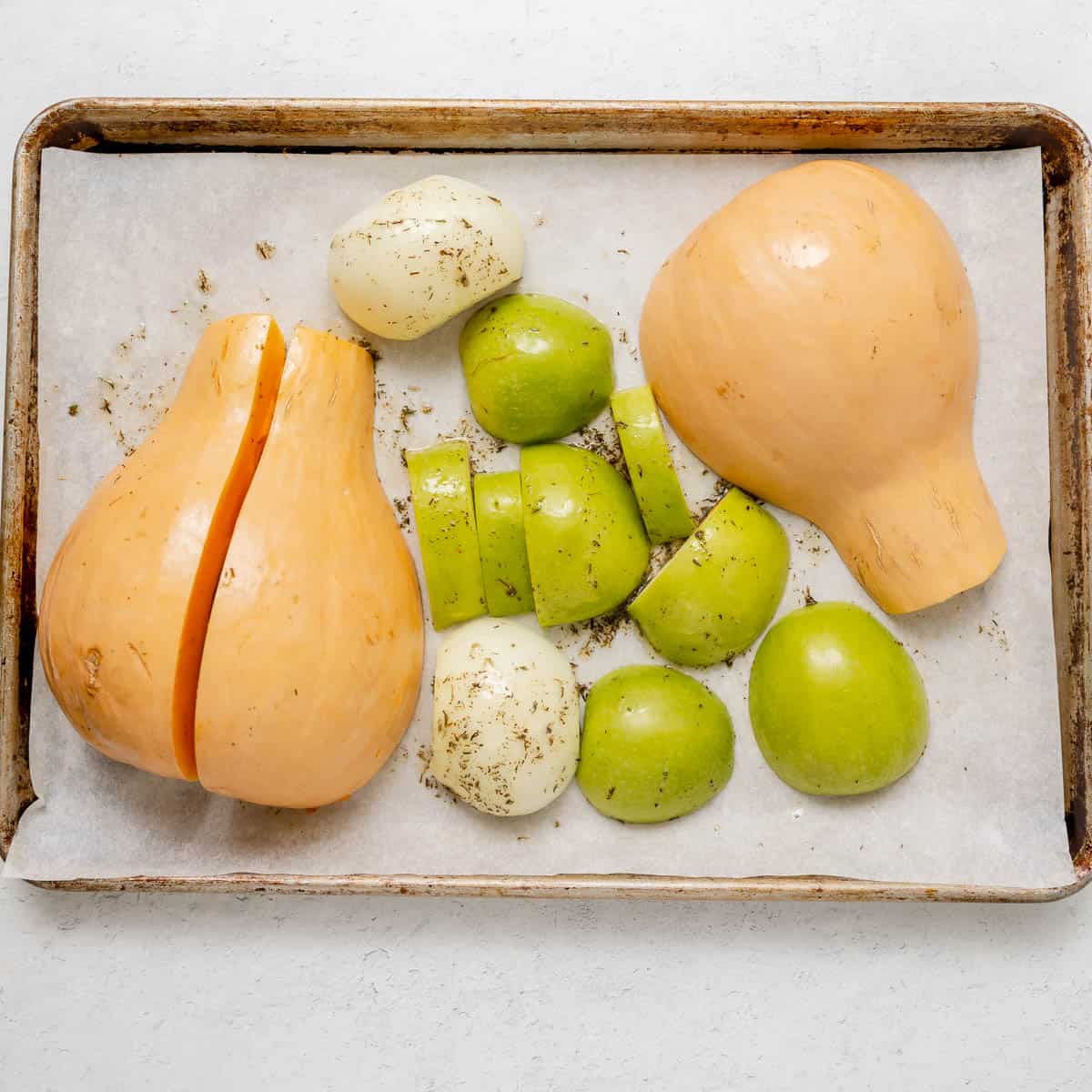Butternut squash halves, green apple pieces, and onion chunks with herbs on a parchment-lined baking sheet.