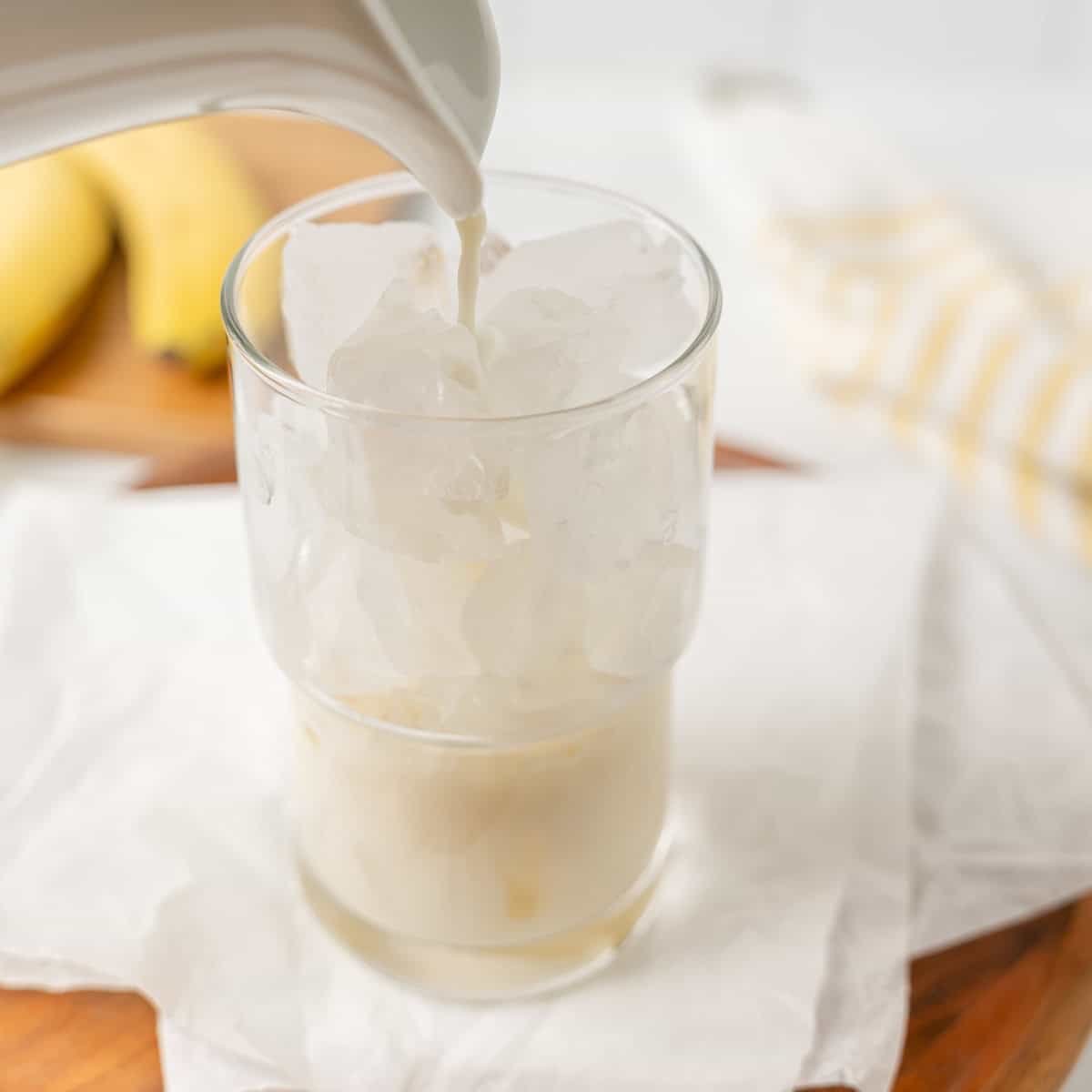 Milk is being poured into a glass filled with ice cubes. Bananas and a striped towel are in the blurred background.