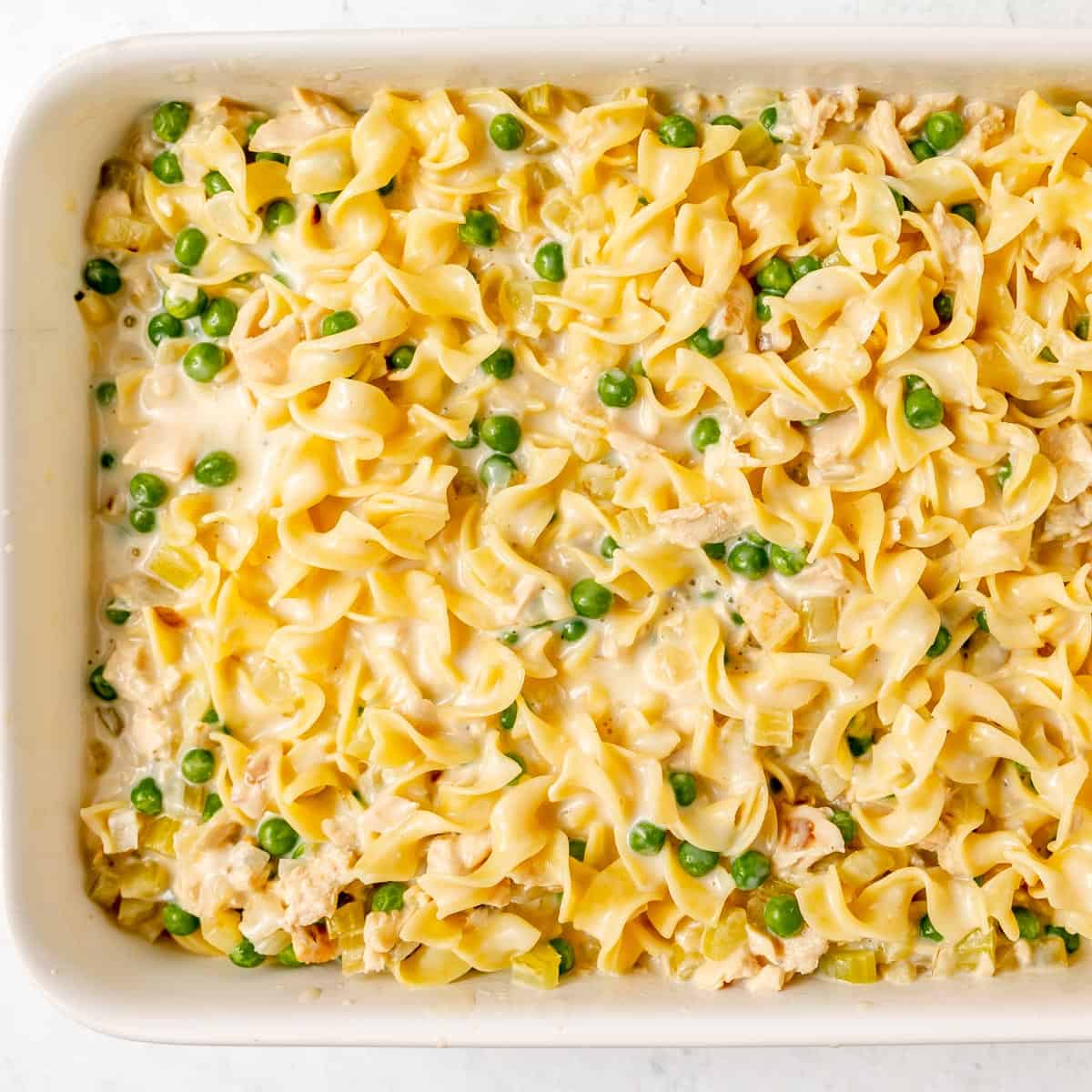 A casserole dish filled with egg noodles, green peas, celery, and a creamy sauce, ready to be baked.