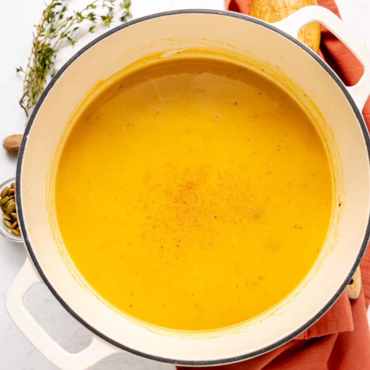 A pot of creamy orange soup sits on a white surface, with a sprig of herbs and a bowl of seeds nearby.