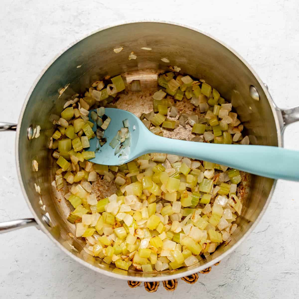 Diced onions and celery are being sautéed in a stainless steel pot with a blue silicone spoon on a light surface.