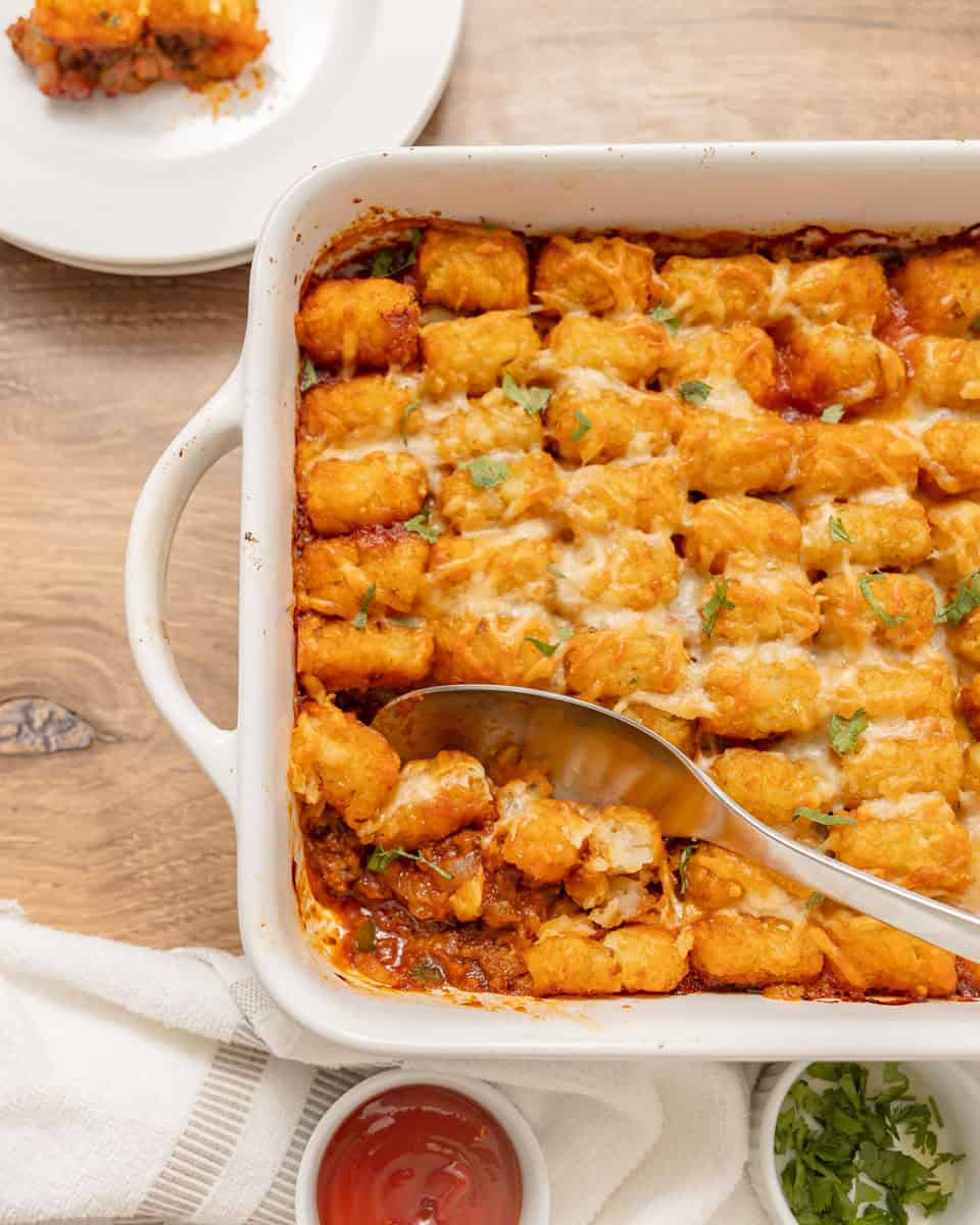 A casserole dish filled with cheesy tater tot casserole, partially served with a large spoon. A plate with a portion, a bowl of ketchup, and chopped herbs are nearby.