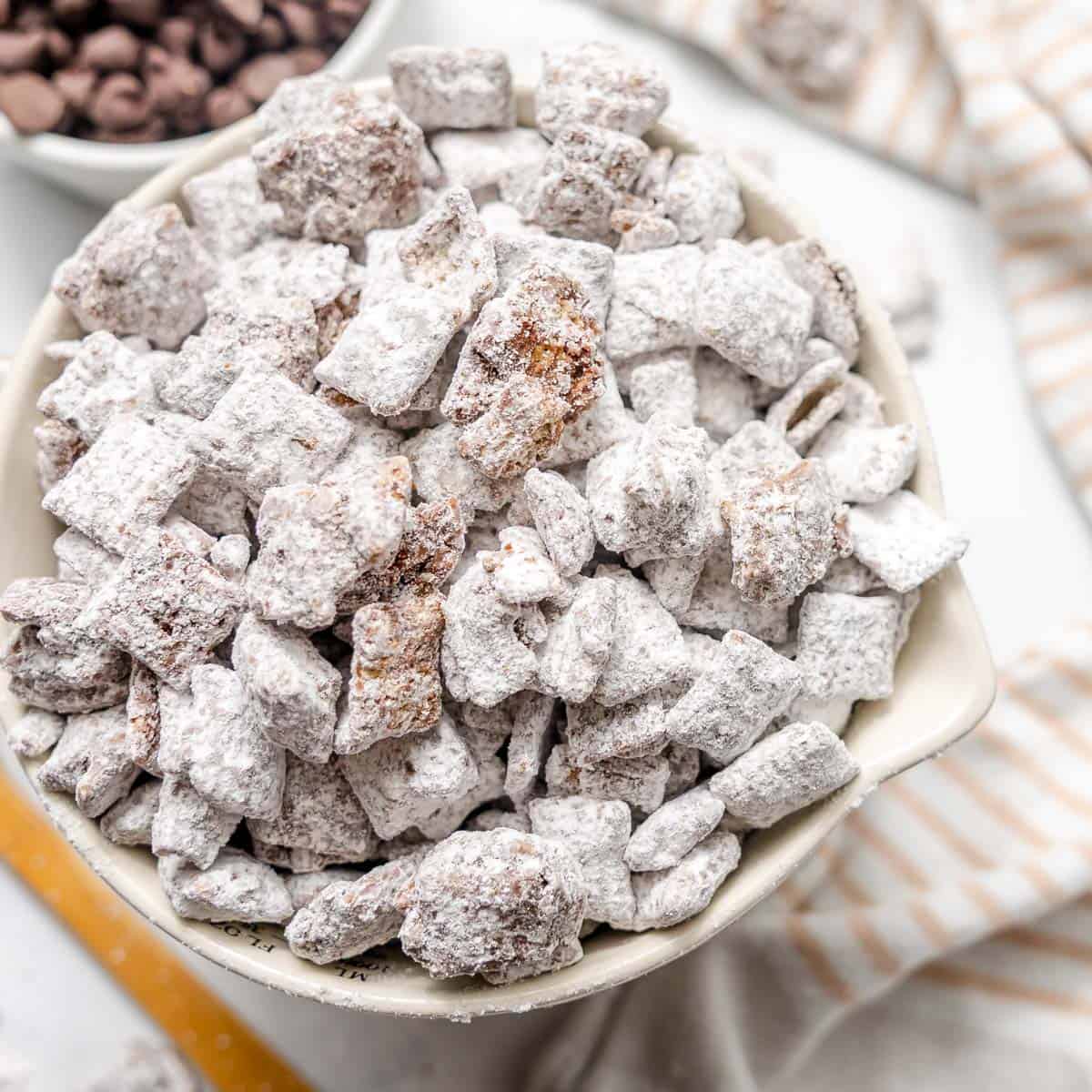 A white bowl filled with powdered sugar-coated snack mix, commonly known as puppy chow or muddy buddies, sits on a white surface.