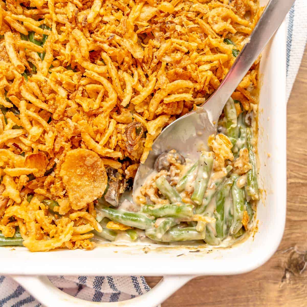A casserole dish of green bean casserole topped with crispy fried onions, partially served with a metal spoon resting inside.