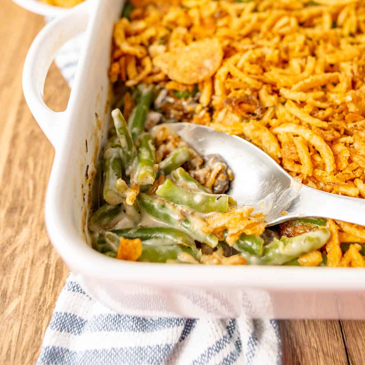 Green bean casserole with crispy fried onions on top, served in a white baking dish with a metal serving spoon on a striped cloth.