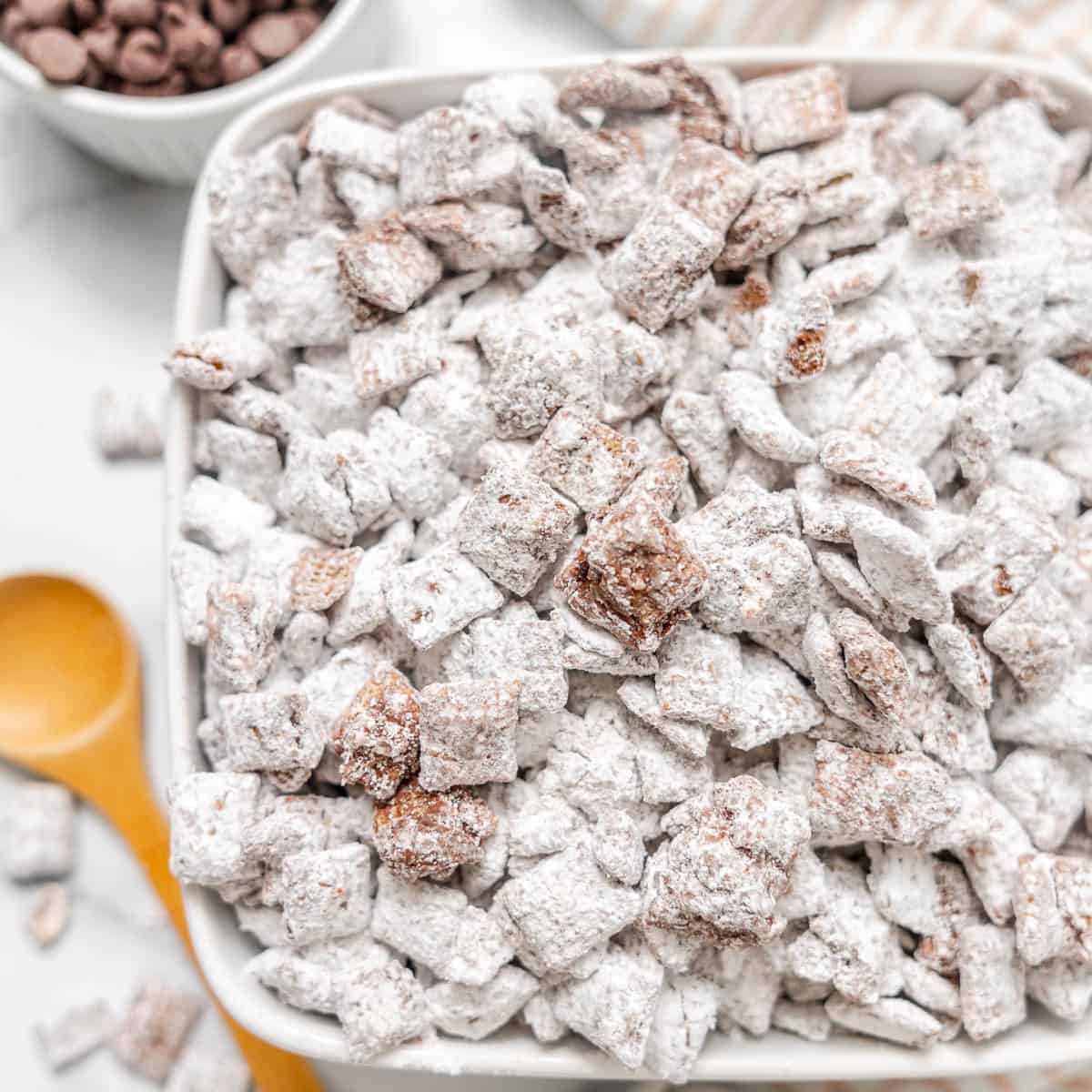 A bowl filled with powdered sugar-coated Chex mix cereal, also known as puppy chow, sits on a white surface next to a wooden spoon and a small bowl of chocolate chips.