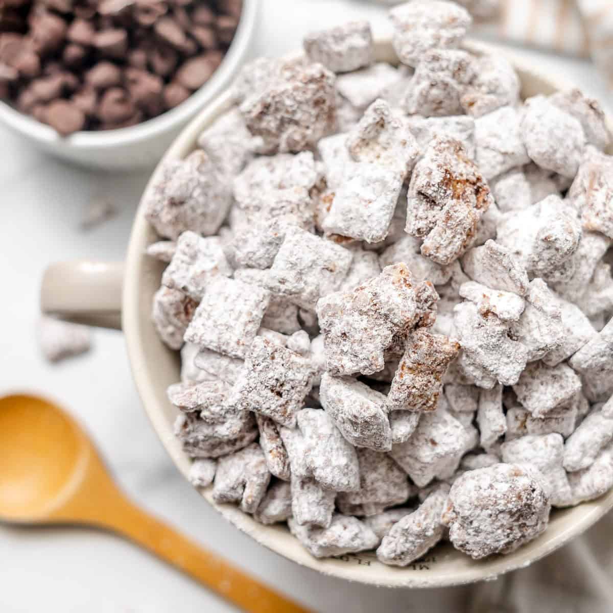 A bowl of powdered sugar-coated snack mix, with a wooden spoon and a bowl of chocolate chips in the background.
