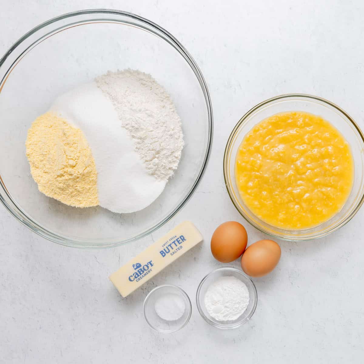 Ingredients for baking are arranged on a white surface: flour, cornmeal, sugar, baking powder, salt, eggs, melted butter, and creamed corn in separate bowls and containers.
