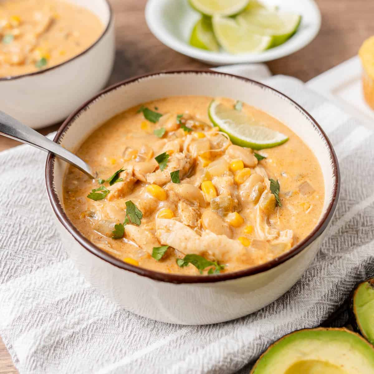 A bowl of creamy chicken and white bean chili garnished with cilantro and a lime wedge sits on a cloth napkin, with avocado slices and lime wedges nearby.