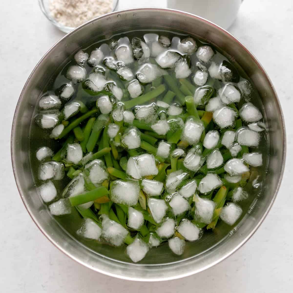 Metal bowl filled with cut green beans and ice cubes submerged in water, on a white surface with a small bowl of flour partially visible in the background.