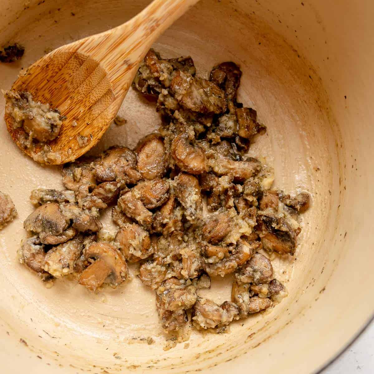 A wooden spoon stirs sliced, browned mushrooms sauteed with herbs and garlic in a light-colored Dutch oven.