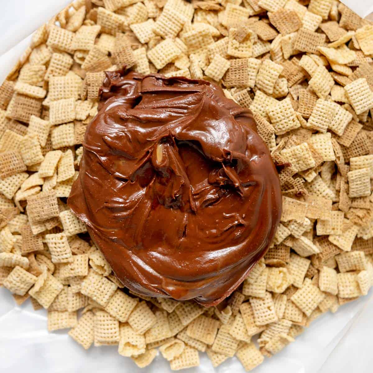 A pile of plain Chex cereal squares with a large dollop of chocolate spread on top, on a white surface.