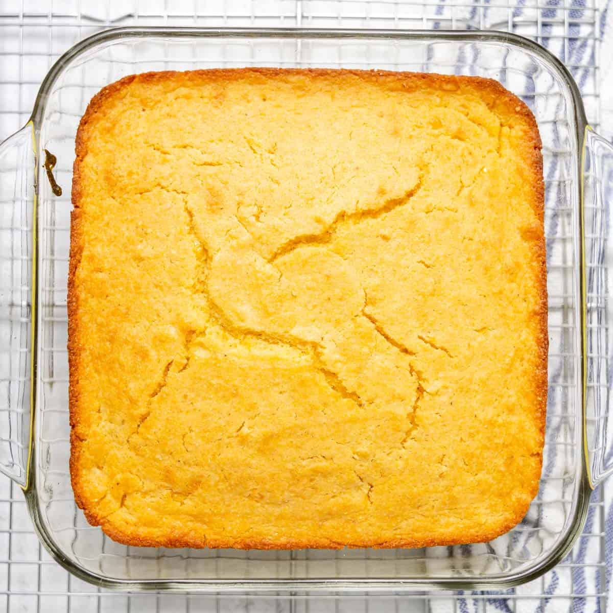 Golden baked cornbread with a cracked top, in a glass square dish, sitting on a wire cooling rack over a white background.