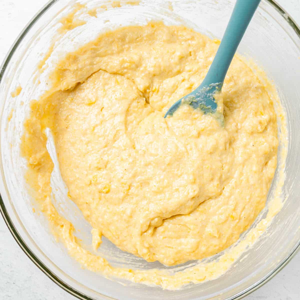 A blue spatula mixes thick, yellow batter in a clear glass bowl on a white surface.