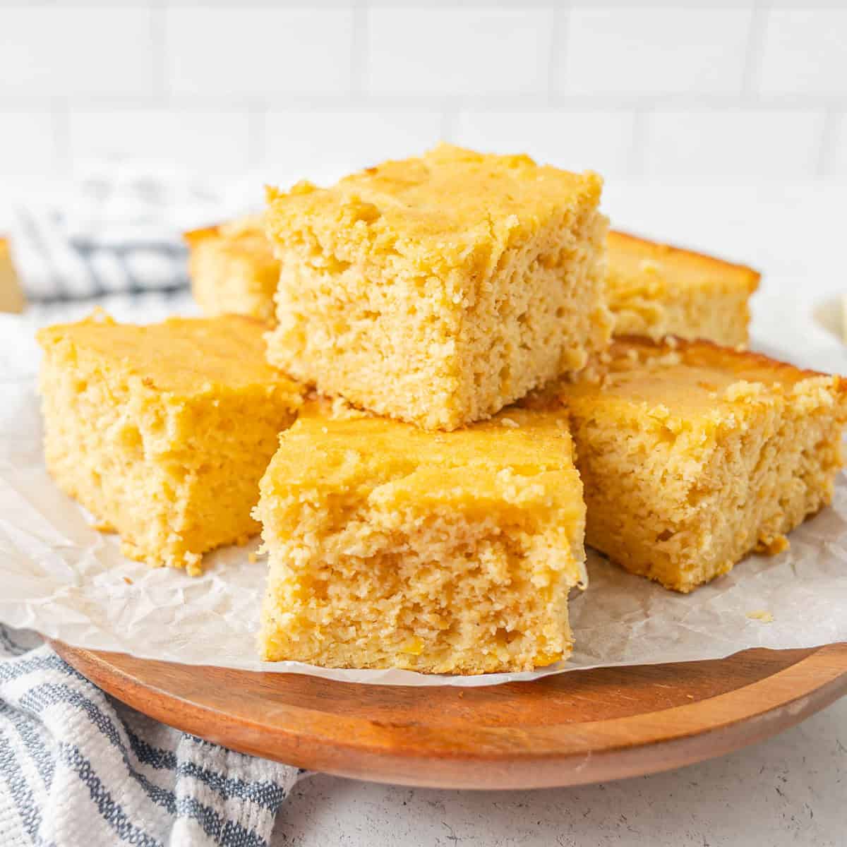 Six pieces of cornbread are stacked on a round wooden board lined with parchment paper, with a white tile background.