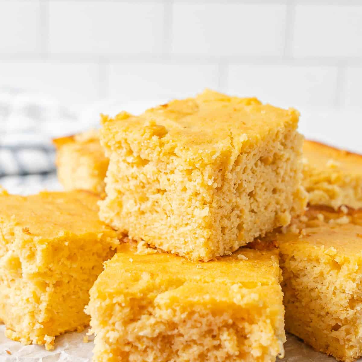 Several pieces of golden, moist cornbread are stacked on a white surface with a tiled background.