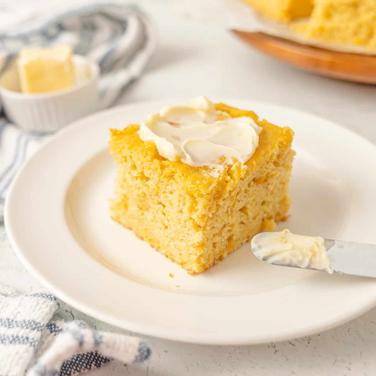 A square piece of cornbread with butter spread on top sits on a white plate, with a butter knife resting beside it.