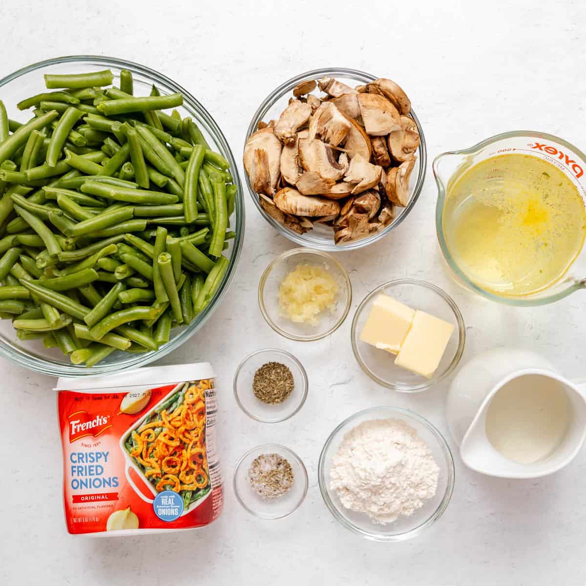 Top-down view of green beans, sliced mushrooms, melted butter, minced garlic, milk, flour, butter, spices, and a container of crispy fried onions arranged on a white surface.