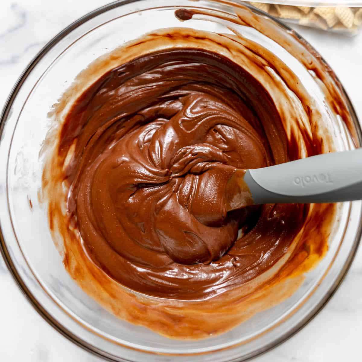 A glass bowl filled with chocolate batter being mixed with a gray spatula on a white surface.