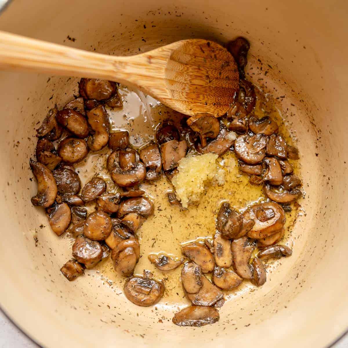 Sautéed sliced mushrooms and minced garlic in oil in a pot, with a wooden spoon resting inside.