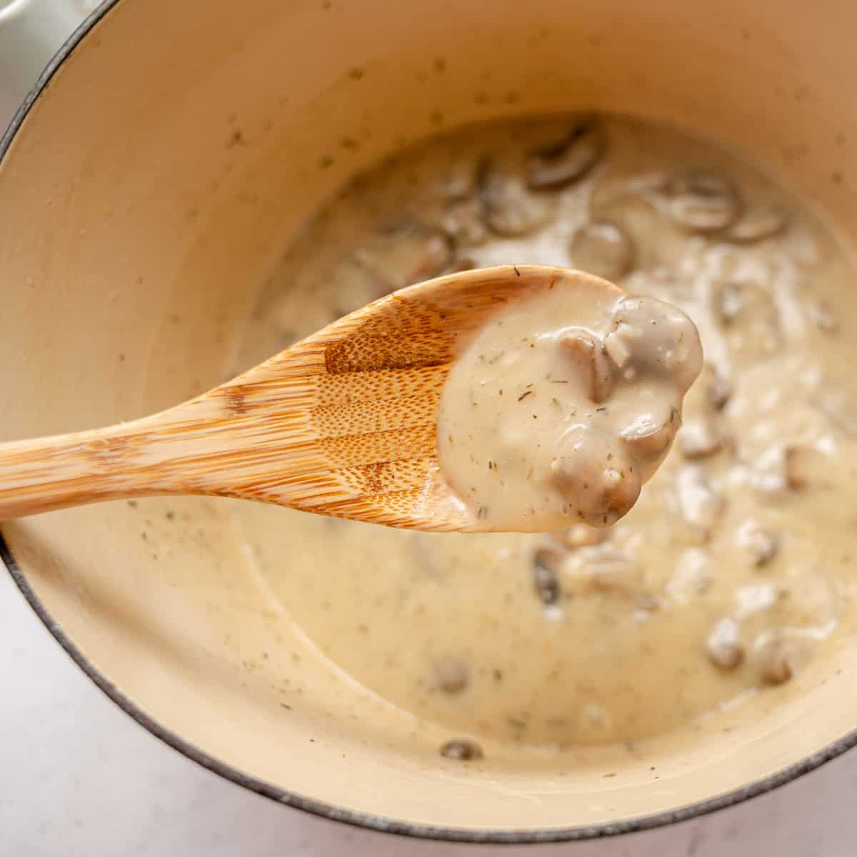 A wooden spoon holding creamy mushroom soup above a pot filled with the same soup.