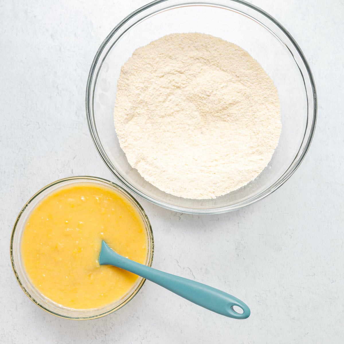 Two glass bowls: one with dry flour mixture, one with wet yellow batter and a blue spatula, on a white surface.