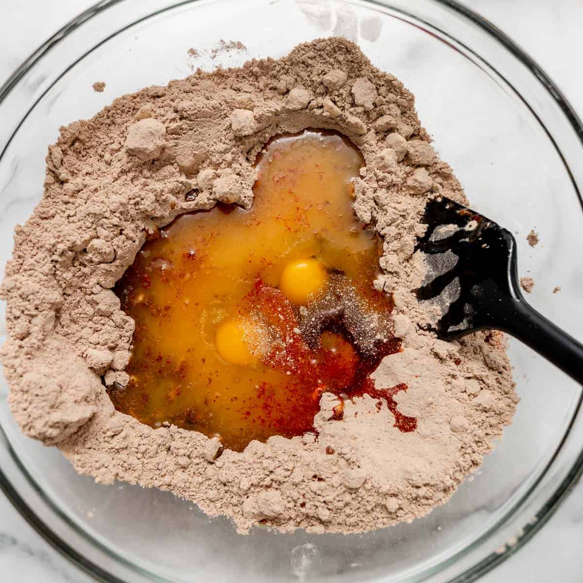 Two raw eggs and liquid ingredients sitting in a well of red velvet cake mix in a glass bowl with a spatula.