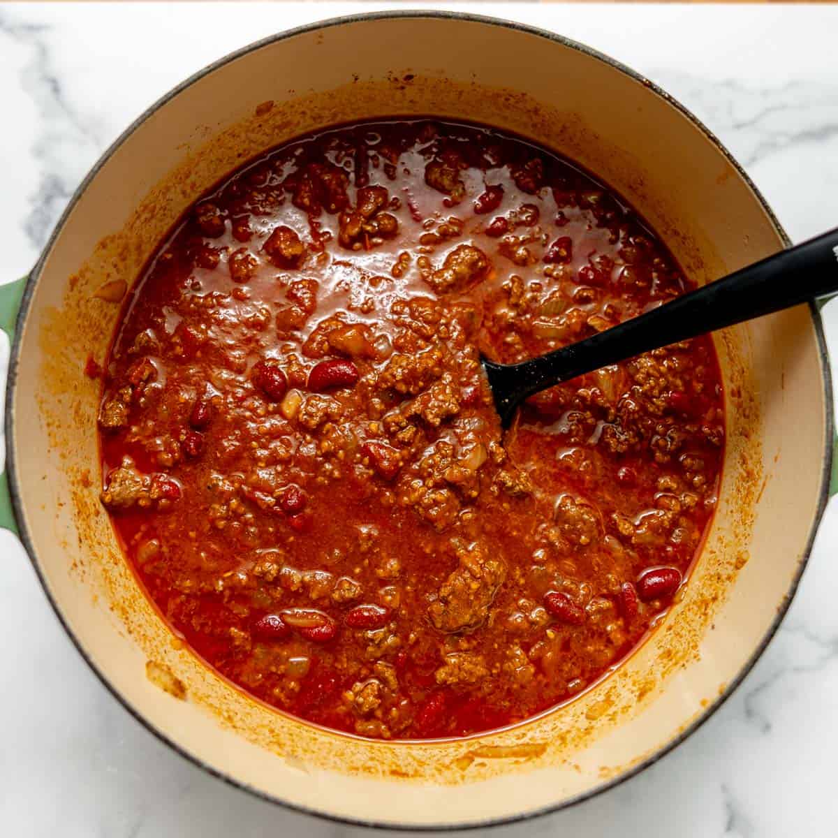 A pot of chili with ground beef, kidney beans, and tomato sauce, being stirred with a black spoon on a white marble surface.