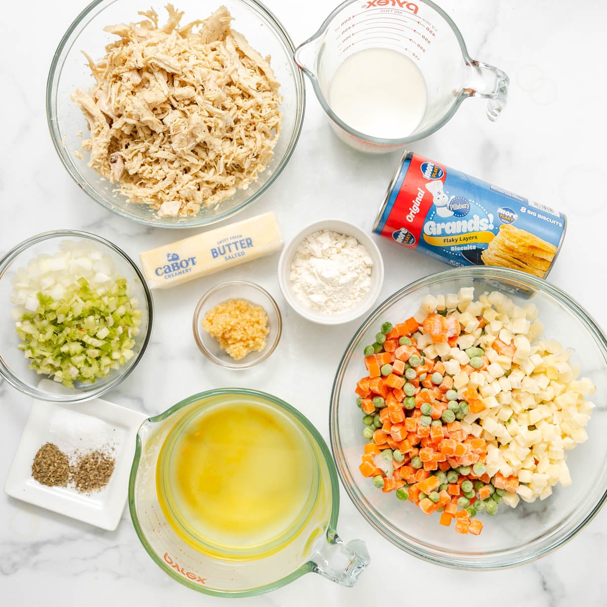 Ingredients for chicken pot pie, including shredded chicken, frozen vegetables, broth, milk, a stick of butter, minced garlic, flour, seasonings, and a tube of refrigerated biscuit dough.