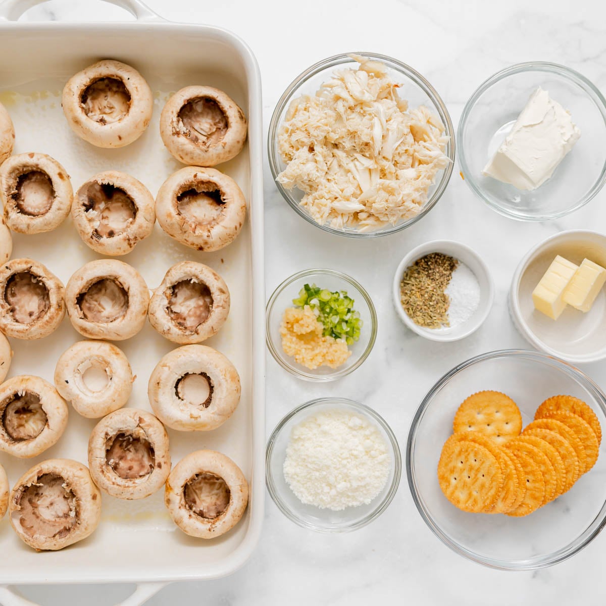 Ingredients for stuffed mushrooms, including mushroom caps in a baking dish, crab meat, cream cheese, butter, spices, cheese, and crackers in bowls.
