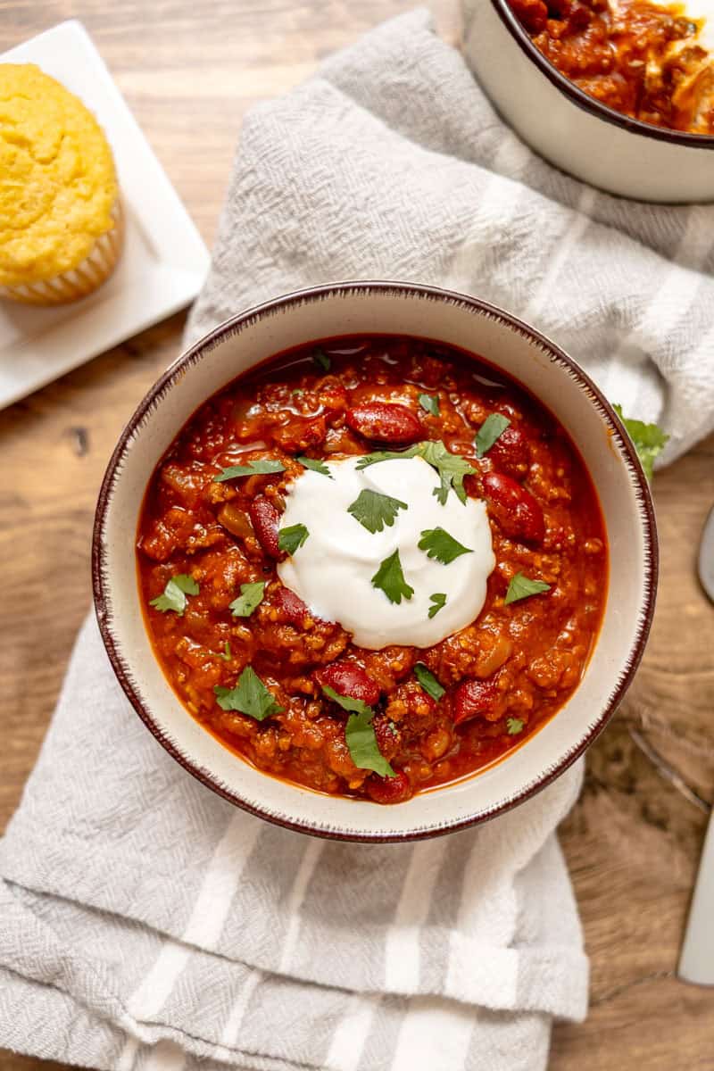 A bowl of chili topped with sour cream and chopped cilantro, on a striped towel, with a plate of cornbread in the background.