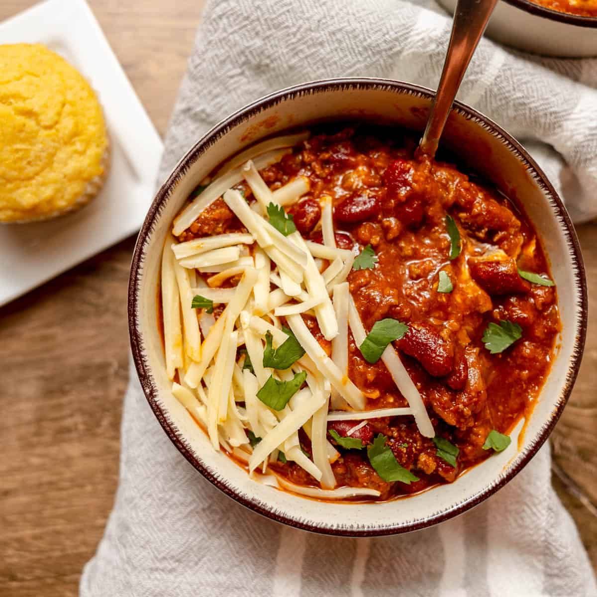 A bowl of chili topped with shredded cheese and parsley sits on a striped cloth, with a piece of cornbread on a small square plate nearby.