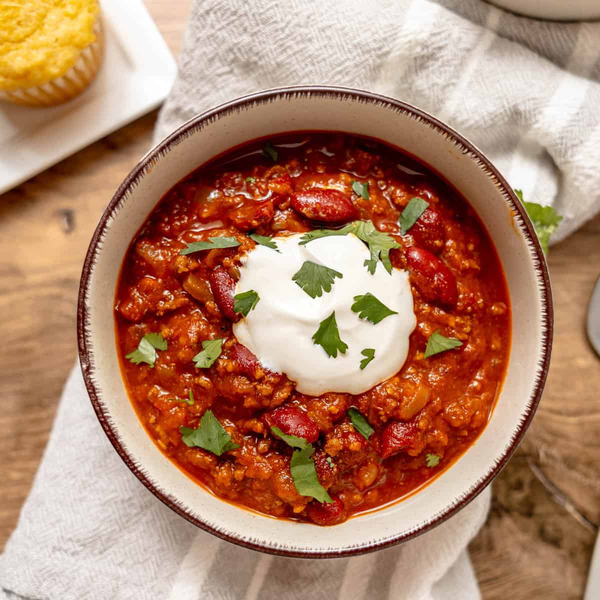 A bowl of chili topped with sour cream and chopped cilantro sits on a cloth napkin, with a piece of cornbread on a plate nearby.